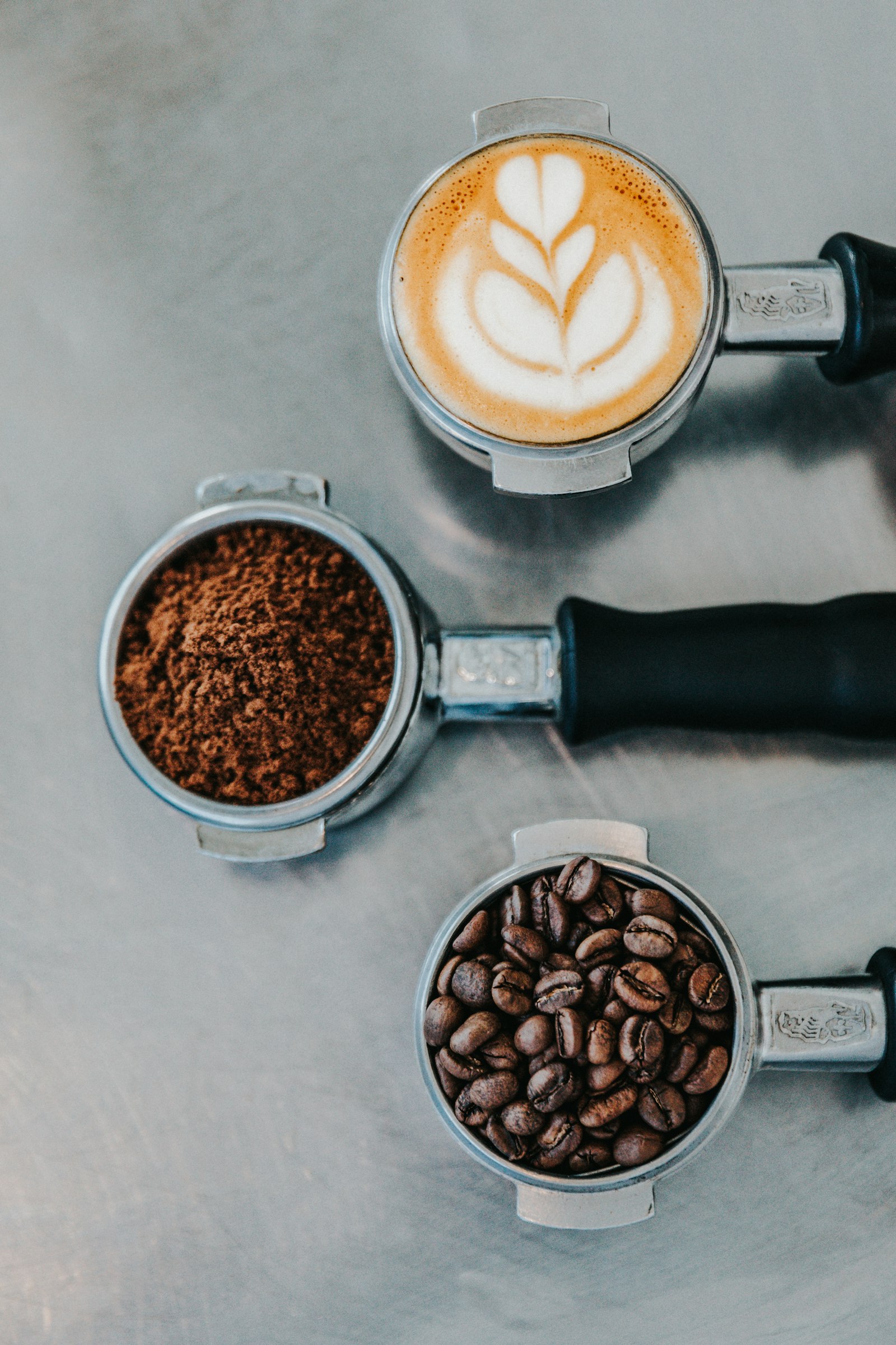 Three espresso portafilters showing coffee beans, ground coffee and a finished latte